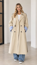 Front full-length view of woman wearing beige double-breasted maxi trench coat with blue contrast cuffs