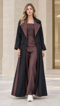 Front full-length view of model wearing black maxi flared trench coat with brown contrast lapel